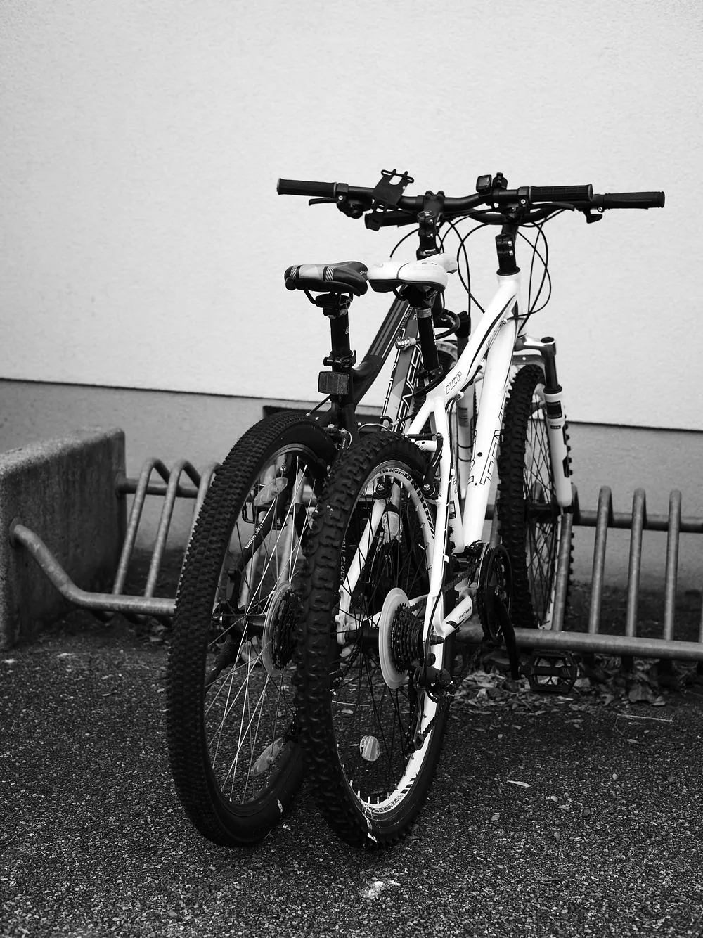 Two mountain bikes parked at a rack against a plain wall, black and white.