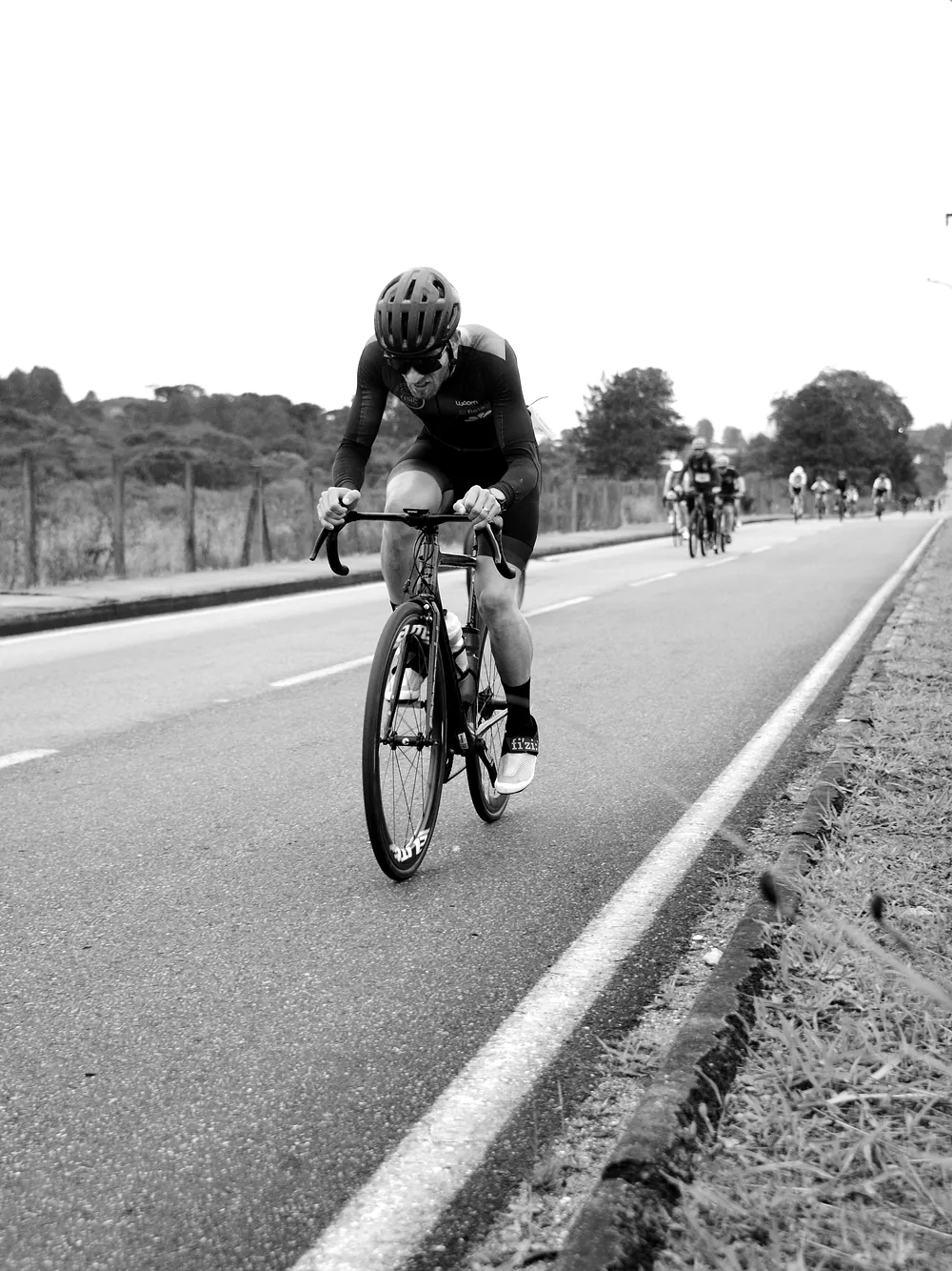 A road cyclist riding toward the camera on an open road with a group behind, black and white.